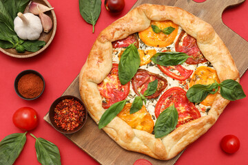 Wooden board of tasty tomato galette with basil and spices on red background