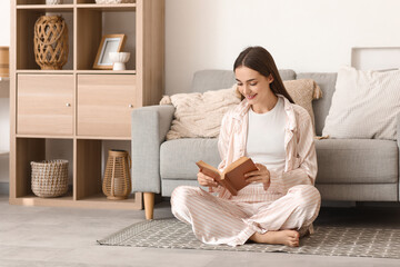 Beautiful young happy woman in pajamas reading book on floor at home