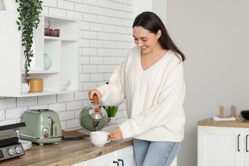 Beautiful young happy woman pouring hot water from modern electric kettle into cup in kitchen