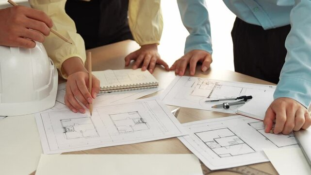 Smart architect and safety engineer discussion about building blueprint on table with architectural document, measurement tools and wooden block scatter around at modern office. Closeup. Delineation.