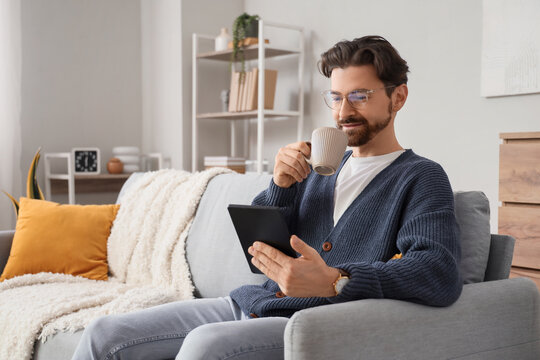Young man with cup of tea and e-reader sitting on sofa at home