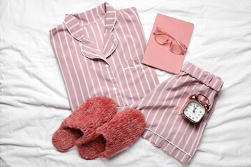 Stylish pajamas with slippers, book and alarm clock on white bed sheet