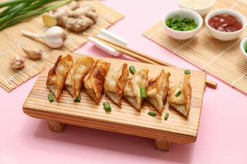 Wooden stand with fried Japanese gyoza, green onion and bowl of sauce on pink background