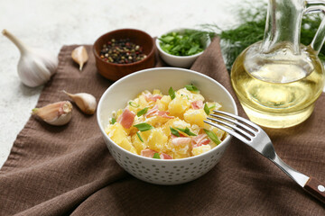 Bowl of tasty potato salad with greens, bacon, garlic and jug of oil on light grunge background
