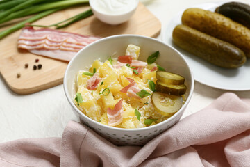 Bowl of tasty potato salad with bacon, green onion and pickled cucumber slices on white background