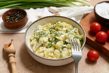 Bowl of tasty potato salad with dill, tomatoes, peppercorns and garlic on beige grunge background