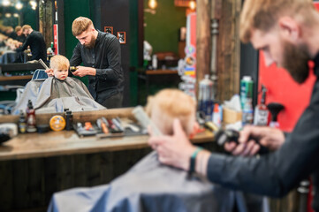 Adorable boy excited about his new haircut. Little client looking at himself in mirror. Reflection in mirror of hairdresser making stylish haircut for his cute client.