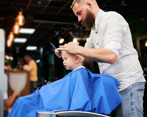 Man barber cutting little boy's hair using comb and scissors. Child getting haircut from adult male, likely barber. Professional hairdresser and cute client at modern barbershop.
