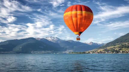Fototapeta premium A vibrant orange hot air balloon floats over a serene lake surrounded by majestic mountains under a bright blue sky.