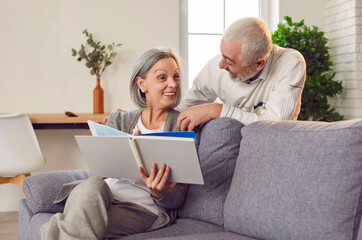 Portrait of a happy cheerful senior elderly couple looking through a family photo album sitting on sofa in living room at home. Mature married man and woman enjoying leisure time together.