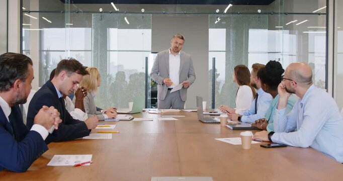 Team of diverse business people having a work meeting in the boardroom, sitting around the office table, listening to a male speaker making a presentation and taking notes on new marketing strategies