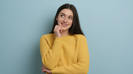 Fototapeta premium Young Woman with Long Dark Hair in Yellow Sweater, Resting Her Chin on Her Hand, Displaying a Cheerful Expression Against a Light Blue Background, Evoking a Relaxed and Thoughtful Mood.
