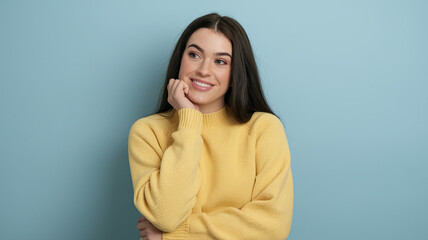 Young Woman with Long Dark Hair in Yellow Sweater, Resting Her Chin on Her Hand, Displaying a Cheerful Expression Against a Light Blue Background, Evoking a Relaxed and Thoughtful Mood.