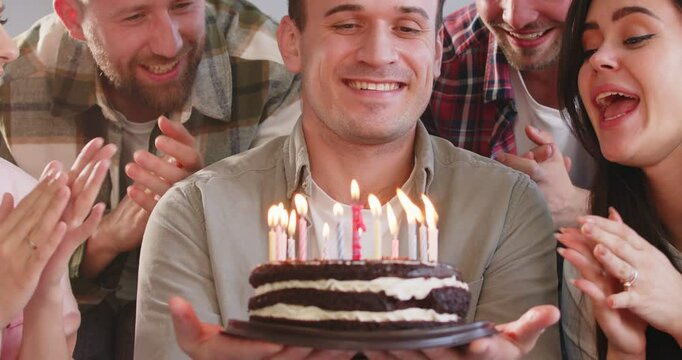 Young happy man blowing out the candles on birthday cake in the living room at home at evening party with best friends congratulating him and clapping hands. Birthday party concept. 4k video.