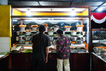 A man serving customers, Indonesian food, Padang cuisine buffet: Fish in coconut milk, fried fish, grilled fish, beef, and other dishes are displayed in a glass display case.