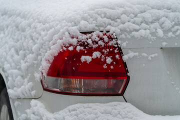 Close-up rear view or elegant red tail light of modern white coloured car covered with fresh white snow outdoors on winter city street. Soft focus. Copy space. Automobile details theme.