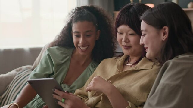 Medium shot of cheerful diverse female zoomers watching digital tablet when sitting on couch during home gathering