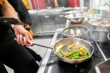 A close-up of a chef's hand skillfully sautéing green beans and golden potatoes in a stainless steel pan in a busy kitchen. Steam rises, showcasing a dynamic cooking environment.