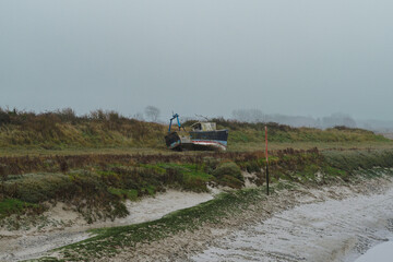 Abandoned fishing boat rests on muddy shore under overcast sky near marshy grasslands
