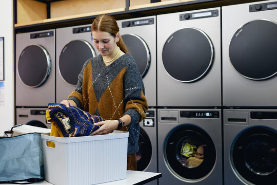 Medium shot of young woman holding vintage sweaters sorting through basket while checking result after washing in laundry machine in coin laundromat, copy space