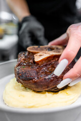 A close-up of a hand placing a perfectly cooked steak with a bone on a bed of creamy mashed potatoes in a modern kitchen setting. The dish showcases gourmet presentation and skillful plating.