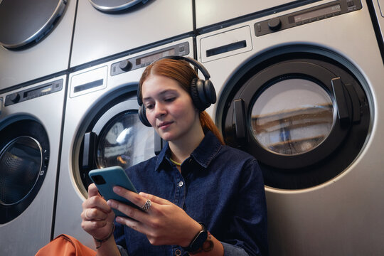 Medium shot of young woman enjoying music in headphones scrolling news feed while waiting in coin laundromat with washing machines tumbling in background - Powered by Adobe
