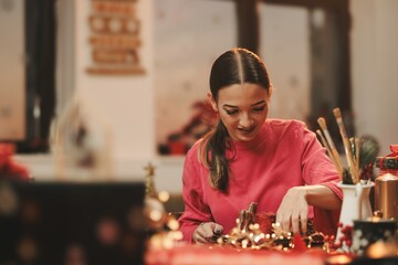 A young woman diligently makes Christmas decorations in a warmly lit workshop. Her focus and joy highlight the festive spirit, surrounded by vibrant colors and creative supplies