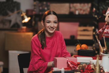 In a warmly decorated room, a cheerful girl smiles while wrapping presents amidst festive decorations, embodying the joy of the Christmas season and holiday spirit
