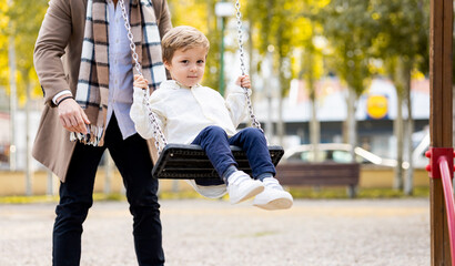 Photograph of a Caucasian father in his 30s pushing his son sitting on a swing. The child looks at the camera while the father is unrecognizable.Concept of parents playing with children in a park.