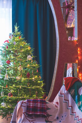 Christmas tree in the corner of the room. There are garlands and glass toys on the Christmas tree. A sofa with a Christmas blanket. A hat and a blanket on the couch.