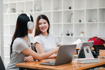 Two young women working together in a modern office setting, using laptops and headphones, engaged in a collaborative discussion.