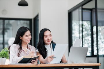 Two businesswomen working together at a desk with laptops and papers in a bright, modern office setting.