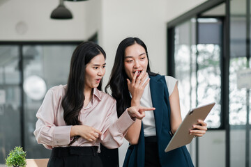Obraz premium Two professional women in a modern office, engaged and excited while looking at a tablet, showcasing teamwork and innovation.
