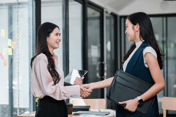 Two businesswomen in a modern office setting shake hands, symbolizing successful collaboration and partnership.