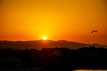Sunset Serenity with Paraglider Silhouette. The glowing sun sets behind distant hills, casting a...