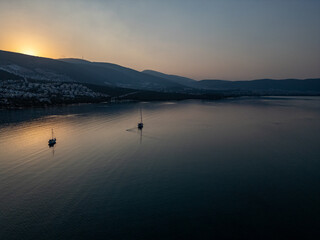 Sunset Serenity on the Bay. Two anchored sailboats rest peacefully on calm waters as the golden hues of sunset illuminate the bay, surrounded by tranquil hills and distant coastal homes.