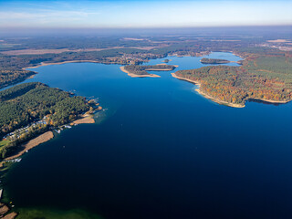 A breathtaking aerial view of a vast lake surrounded by dense autumn-colored forests, dotted with islands and bordered by rural landscapes under a clear blue sky.