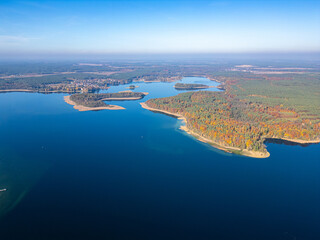 A stunning aerial view of a serene lake surrounded by vibrant autumn forests, small islands, and a peaceful rural town under a bright blue sky.