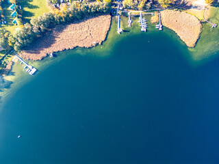 A vibrant aerial top-down view of a lake shoreline with docks, boats, and dense reeds, surrounded by lush greenery and clear blue waters.