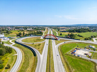 Vibrant Highway Interchange with Overpass. Aerial view of a curved highway interchange featuring a red overpass, surrounded by green fields, rural settlements, and industrial areas under a bright blue
