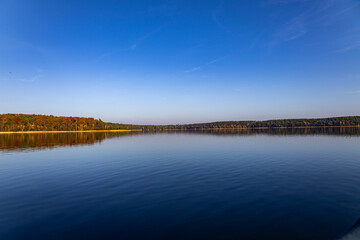 Expansive Autumn Horizon, Expansive lake under a cloudless blue sky with vibrant, warm autumn tones reflecting along the distant treeline, capturing serenity.