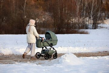 Young woman with a baby stroller walking in winter park. Snow weather, concept of motherhood, single mom with pram