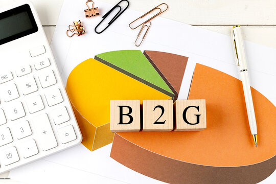 Wooden blocks with the text B2G on a colorful pie chart with a calculator and paper clips on a desk