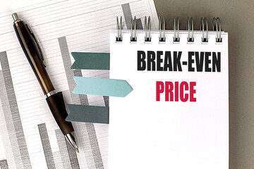 Notebook with the text break-even price, pen, financial charts, and sticky notes on a gray desk