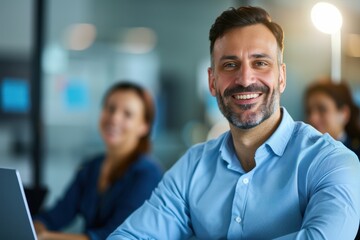 Fototapeta premium A smiling man in a blue shirt sits in an office environment, with colleagues in the background, reflecting a positive and collaborative atmosphere.