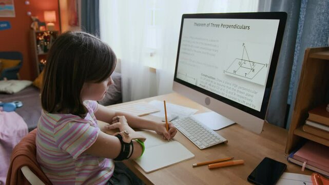 School student with prosthesis looking at computer monitor displaying geometric theorem when taking notes in copybook at wooden desk in bedroom