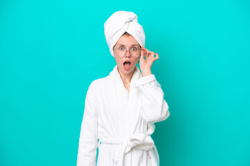 Young woman in a bathrobe isolated on blue background with glasses and surprised