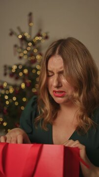 Displeased sad millennial woman opening red present gift box at home. Upset female disappointed with the bad birthday or terrible, horrible, awful anniversary present. 