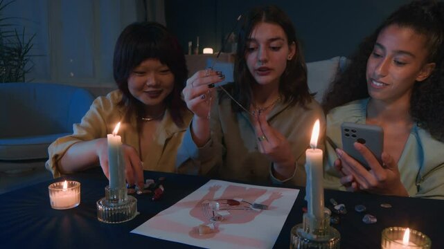 Handheld shot of diverse female zoomers engaged in spiritual ritual using pendulum, crystals, and candles gathering at home during night