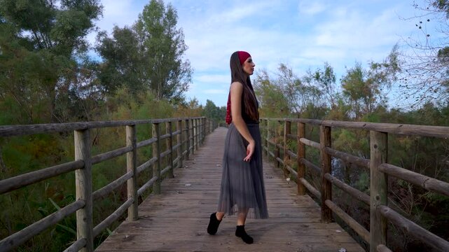 Mujer joven bailando danza contempor&aacute;nea sobre un puente de madera en el bosque al atardecer un d&iacute;a con cielo azul y nubes.